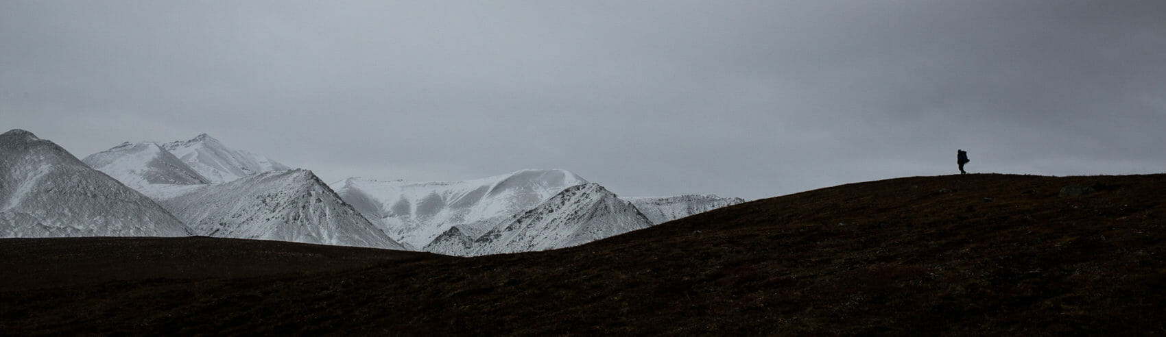 a hiker in the arctic is in silhouette with the snowy brooks range behind